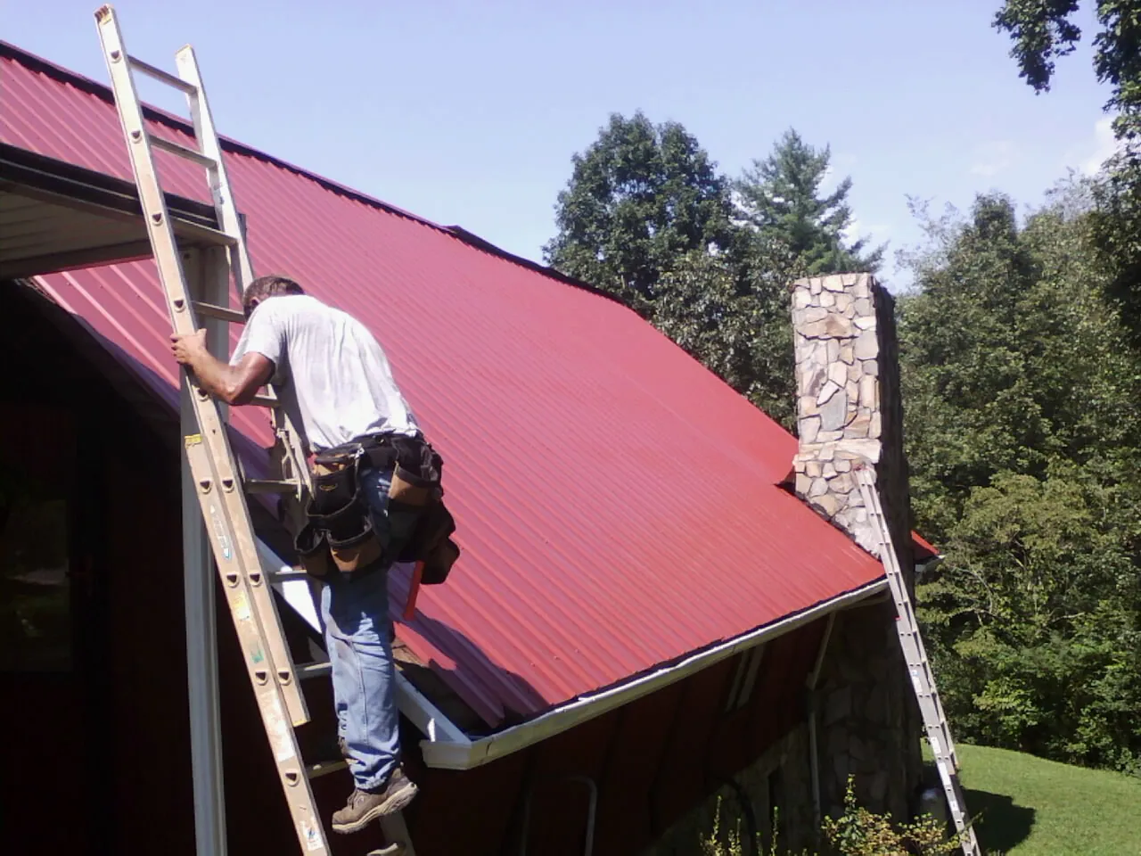Roofer on a ladder beside a red metal roof and stone chimney — Soffit & Fascia Repair by Roofing Brothers in Moundville